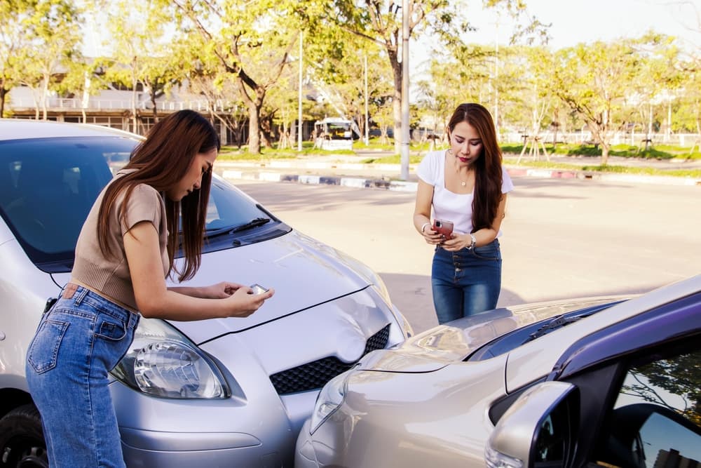 Two women in a car take photos with smartphones after a collision, illustrating accident documentation for insurance claims.