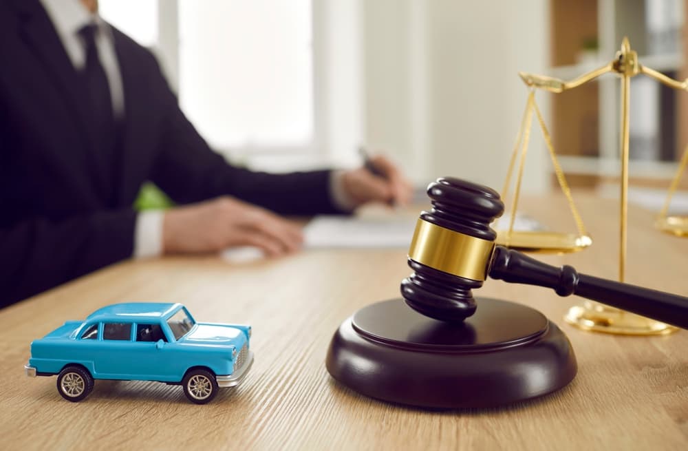 Small toy car and judge's gavel on a table with a judge signing a document in the background—symbolizing a car auction, accident ruling, or insurance claim.