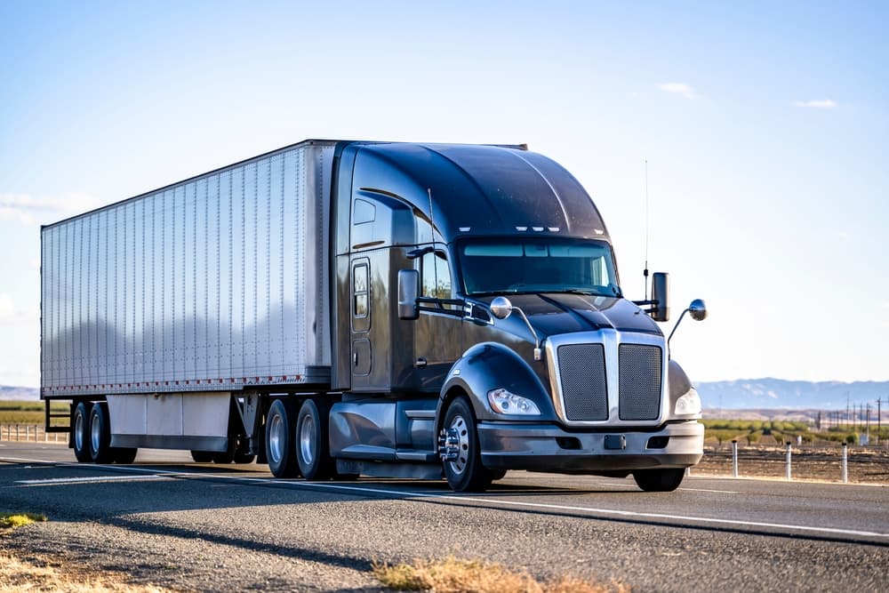 Black big rig semi truck with a sleeper cabin, hauling cargo in a dry van trailer, cruising on the highway.