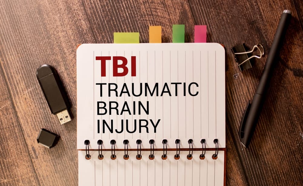 Close-up image of the words 'TBI traumatic brain injury' written on a crisp white notepad against a striking black backdrop.