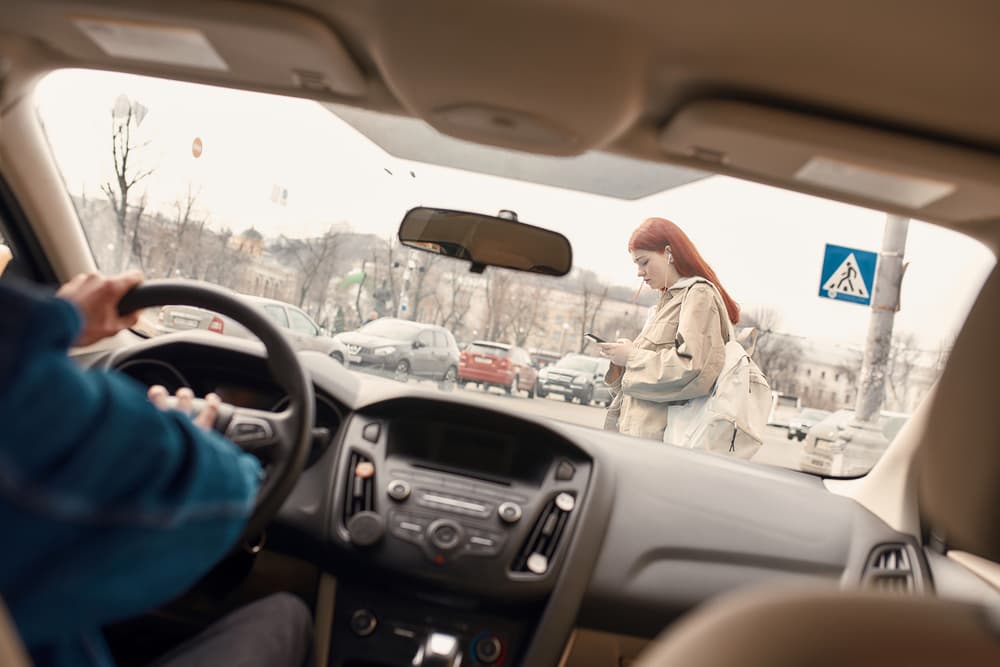 A distracted teenage girl texts on her mobile phone while crossing the street, with a male driver honking the horn in the background. Illustrating the intersection of technology and transportation. Selective focus on the pedestrian.