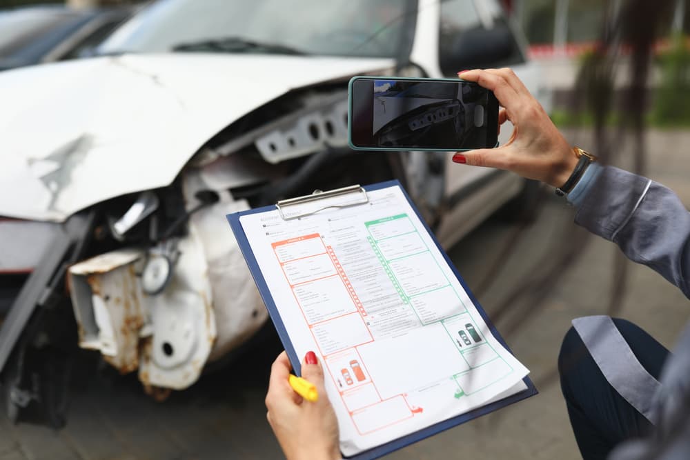 Female taking photos of damaged car with smartphone, holding insurance papers. Car accident assessment.