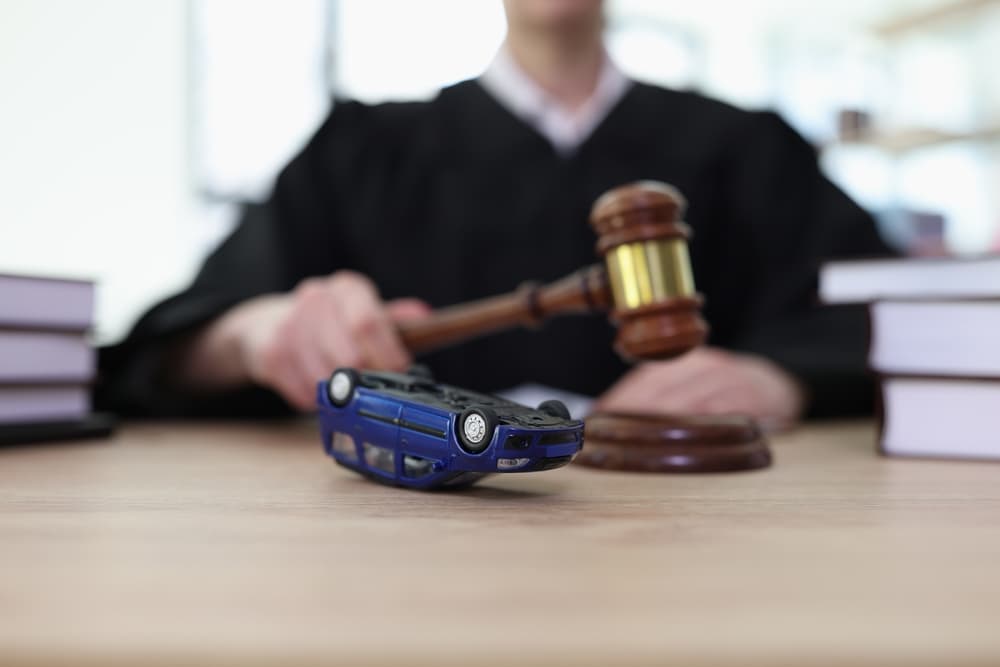 Upside-down toy car beside female judge with gavel in courthouse courtyard. Investigating road accident conditions in court.