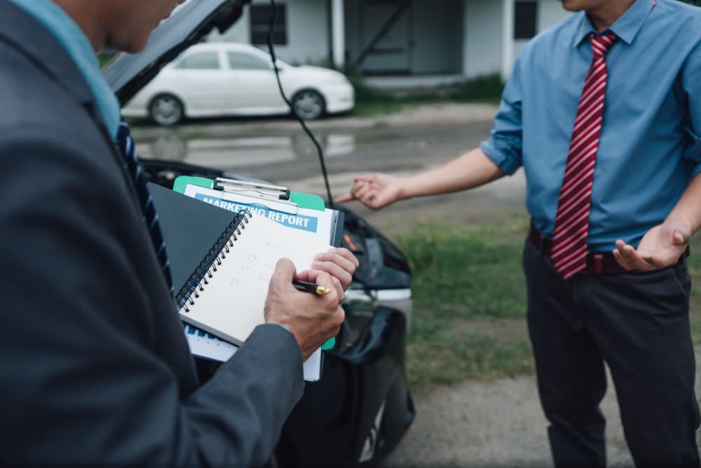 An insurance agent processes a claim payment. They inspect a damaged car as the customer reviews the claim report form.