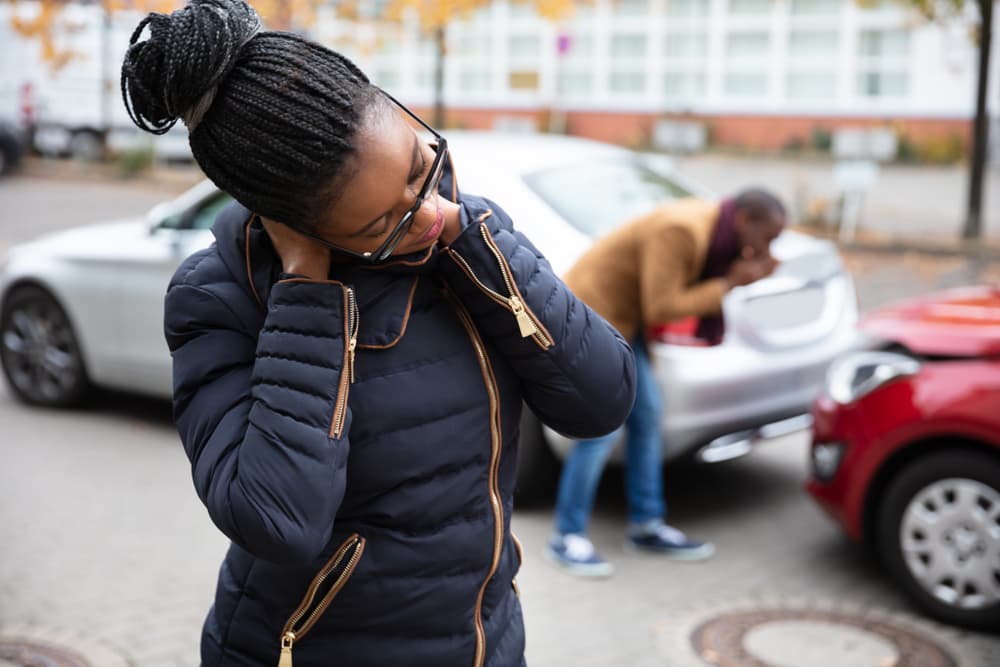 Photo of a woman with neck pain in front of a man inspecting a damaged car on the road after an accident.