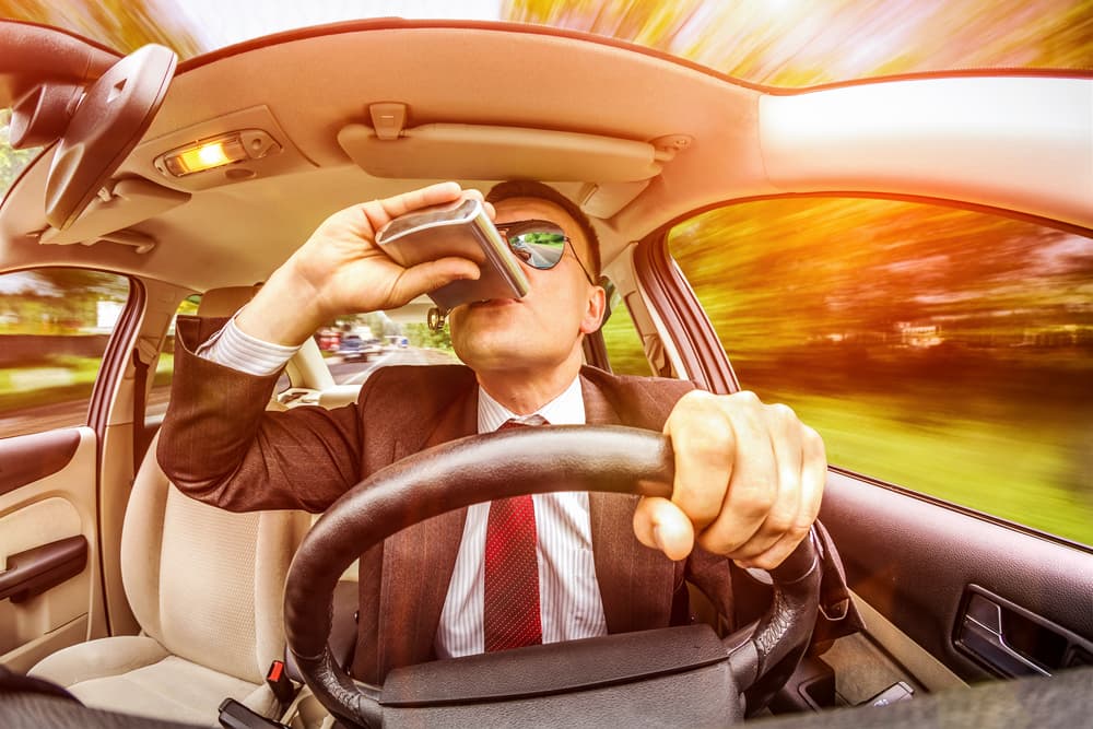 Man wearing sunglasses and a suit driving a car on the road under the influence of alcohol.