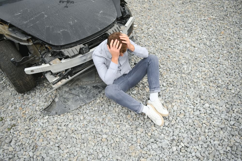 Young Caucasian man involved in a car accident, standing near a damaged vehicle on the road. He holds his head in pain, showing signs of injury and distress.