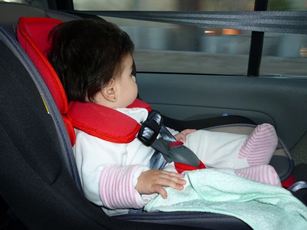 Baby girl secured in a car seat while traveling. Child safety belt fastened for a safe journey. Booster seat, car interior, window view—following traffic rules for protection.