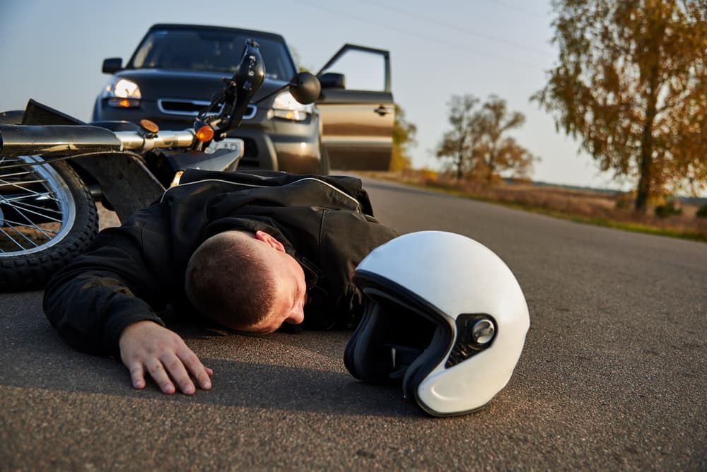 A motorcyclist lies on the pavement beside a motorcycle and car, highlighting the aftermath of a road accident.