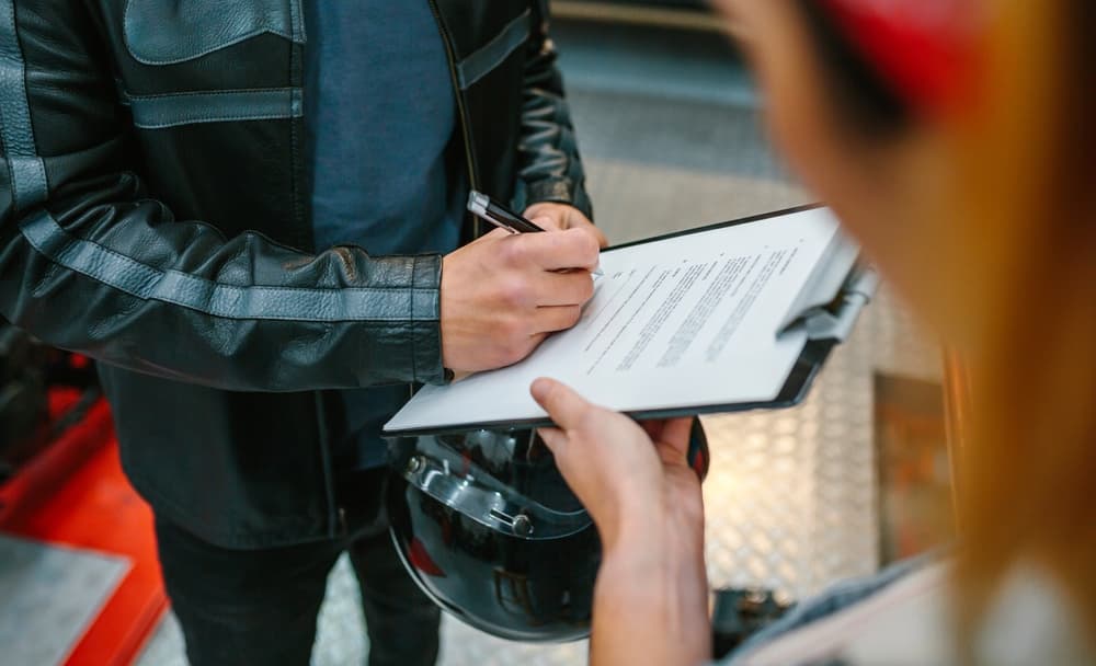 An unrecognizable biker in a leather jacket holds a helmet while signing an insurance document to collect his repaired motorcycle at the workshop.