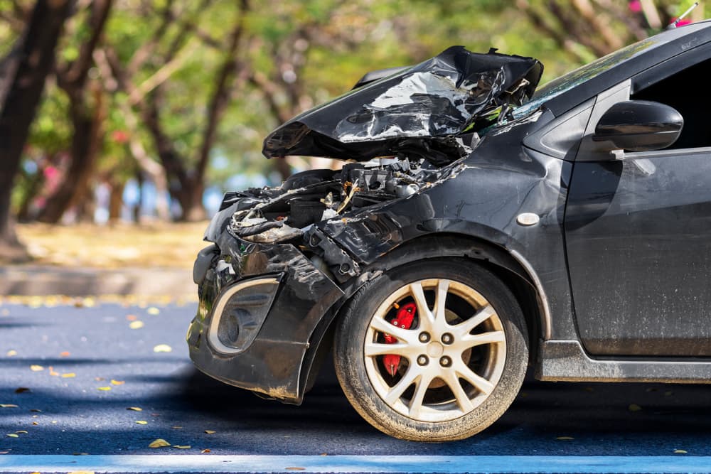 The front of a black car is heavily damaged and broken from a city street accident, parked and no longer drivable. Space available for text or design.