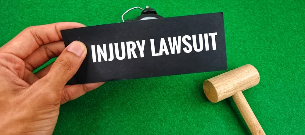 "Injury lawsuit" text displayed on a black card with a wooden gavel placed over a green background – symbolizing legal action, personal injury claims, court justice, and compensation case representation.