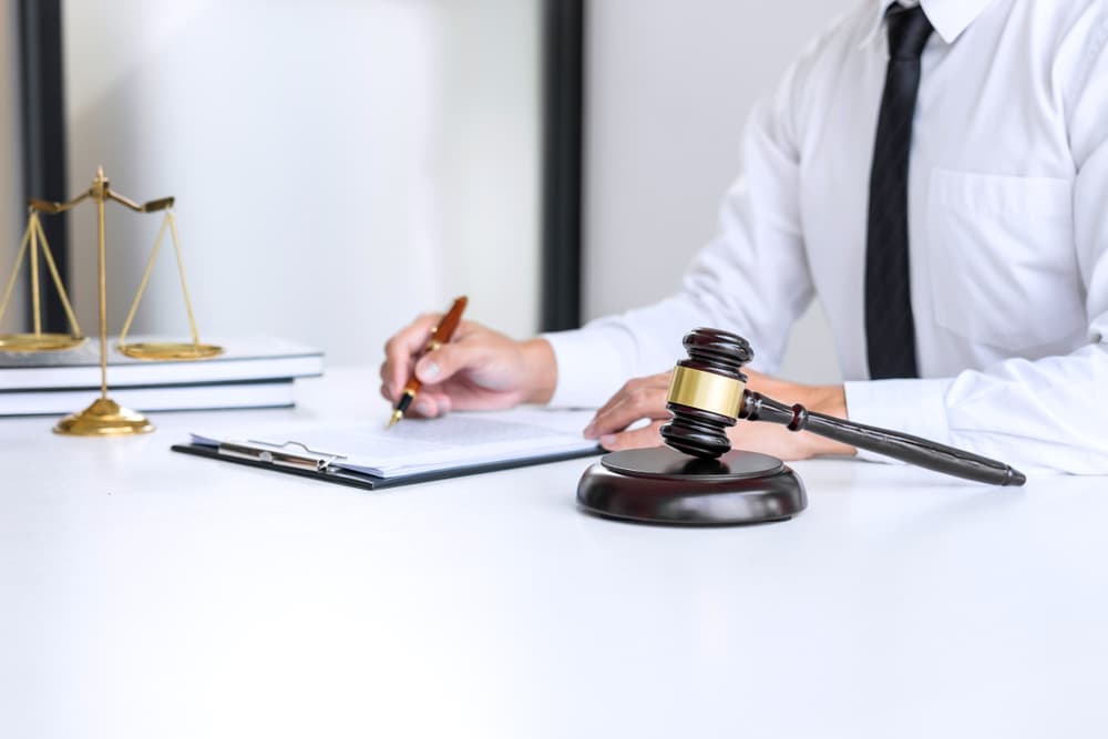 A businessman or attorney reviews legal documents at a desk, with a judge's gavel and justice scales in the background—symbolizing law, legal advice, and justice in a professional law firm setting.