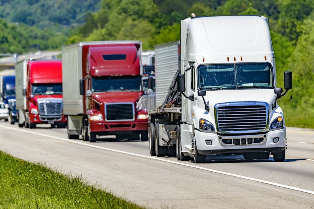 A steady line of eighteen-wheelers speeds down an interstate highway, with heat waves rising from the pavement creating a shimmering effect on the vehicles and trees in the background.